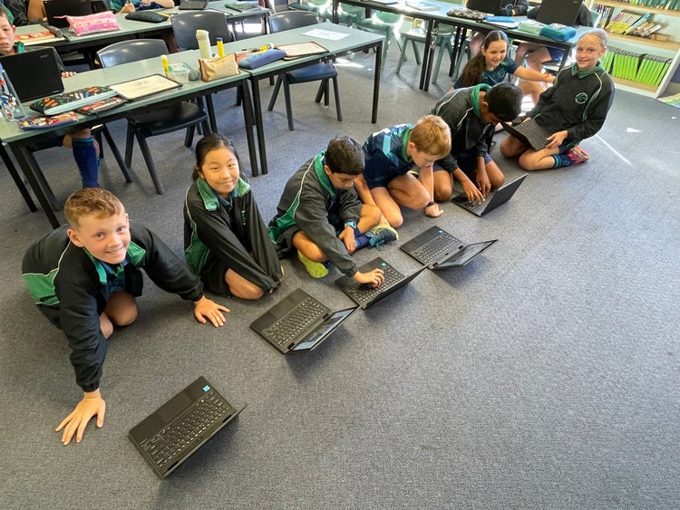 A row of students sitting on the floor in a classroom working on laptops.